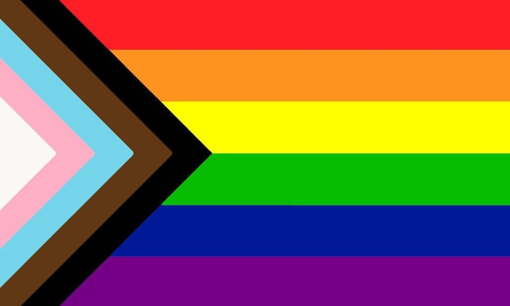 An image of the "progressive pride flag" - on the left side is a white triangle, surrounded by chevrons of pink, light blue, brown, and black. This takes up roughly a third of the flag. The remainder of the flag is covered with six horizontal strips, each a color of the "traditional" pride flag (from top to bottom: red, orange, yellow, green, blue, purple).